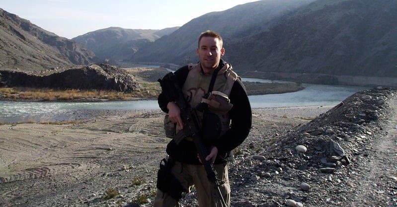 Jason Kander in Afghanistan, standing on a riverbank with mountains behind him